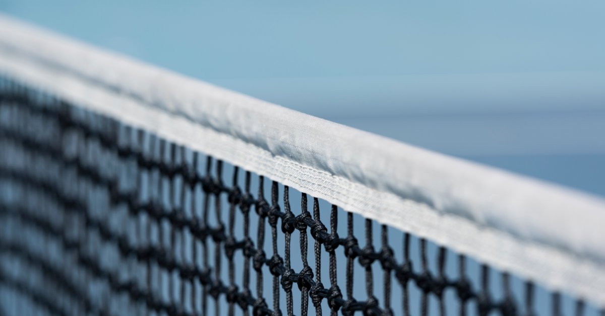 A Guide to Choosing the Right Tennis Net for Your Court A black tennis net with a white top band running diagonally over a blue court, with mesh knots in sharp focus.