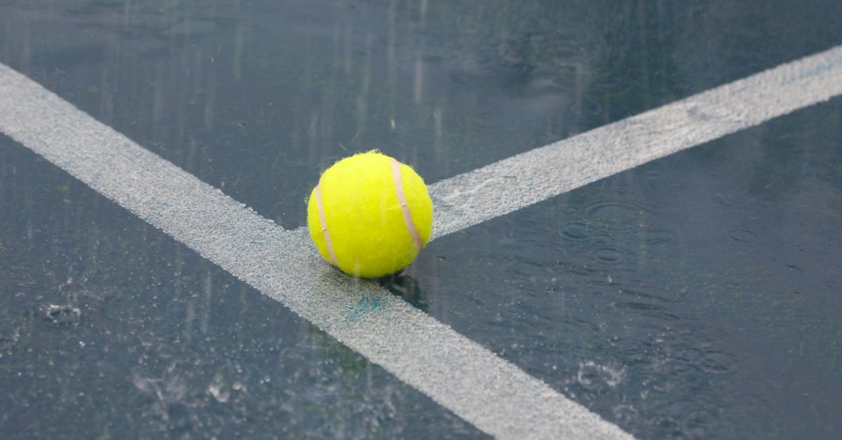 9 Tips for Managing Tennis Rain Delays Effectively A yellow tennis ball resting on intersecting white court lines as rain falls onto the wet blue playing surface.