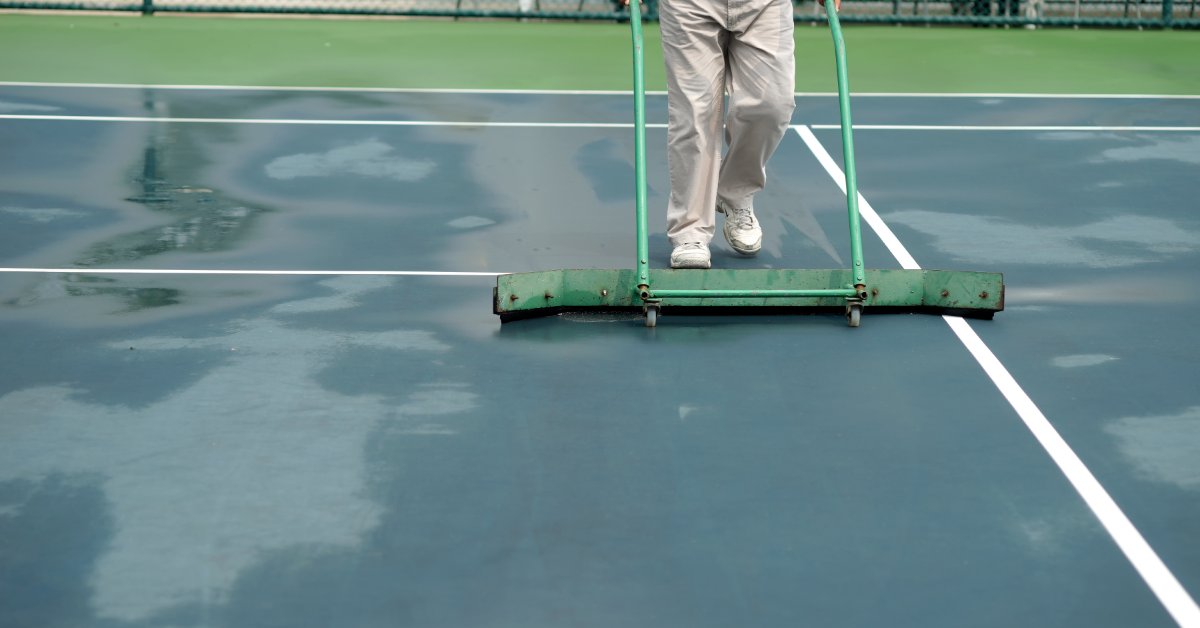 9 Tips for Managing Tennis Rain Delays Effectively A person pushing a green court squeegee across a wet tennis court with puddles and white lines visible.