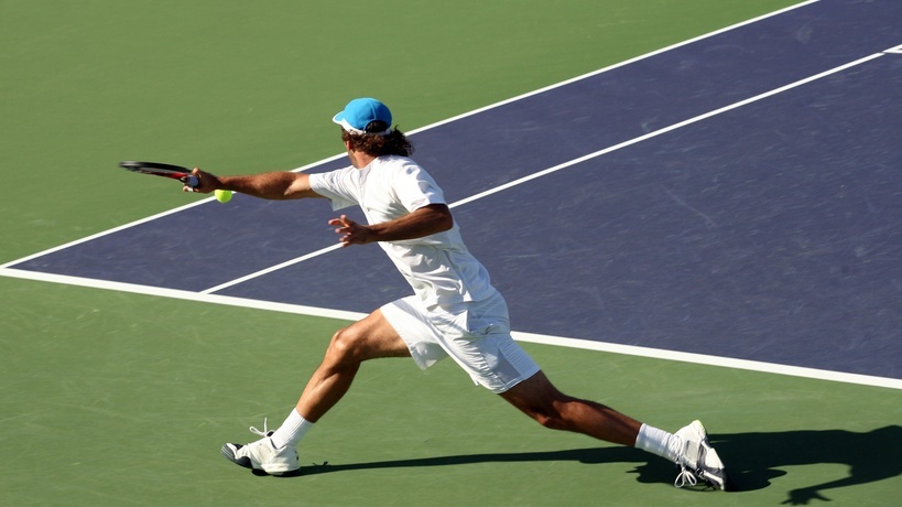 A male tennis player wearing an all-white outfit and a blue hat attempts to hit a backhand stroke with his right hand.