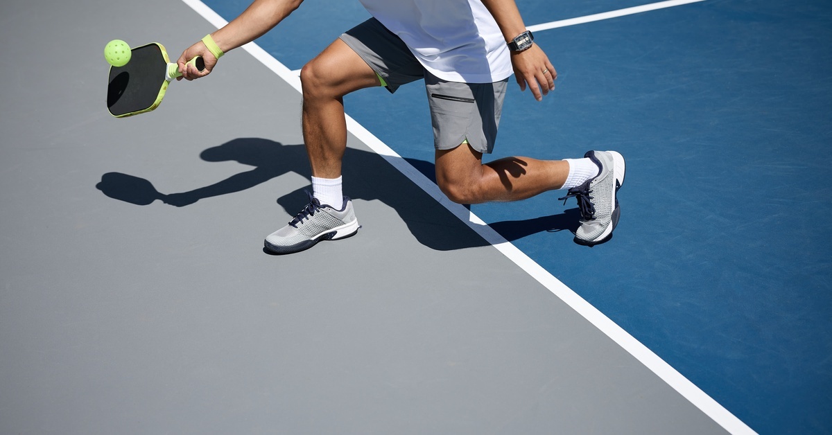 A man wearing a white shirt and gray shorts holds a paddle and hits a pickleball on a blue and gray court.