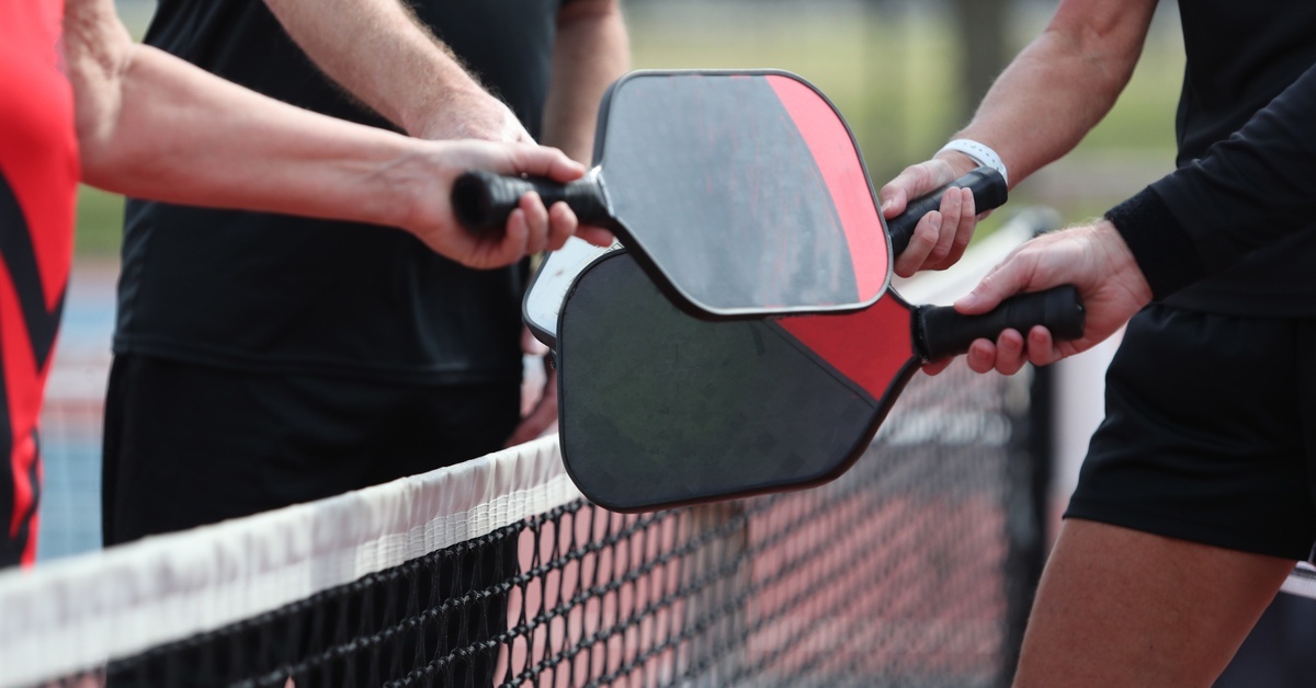 Four people gather around a black net and bump their black-and-red pickleball paddles together during the day.
