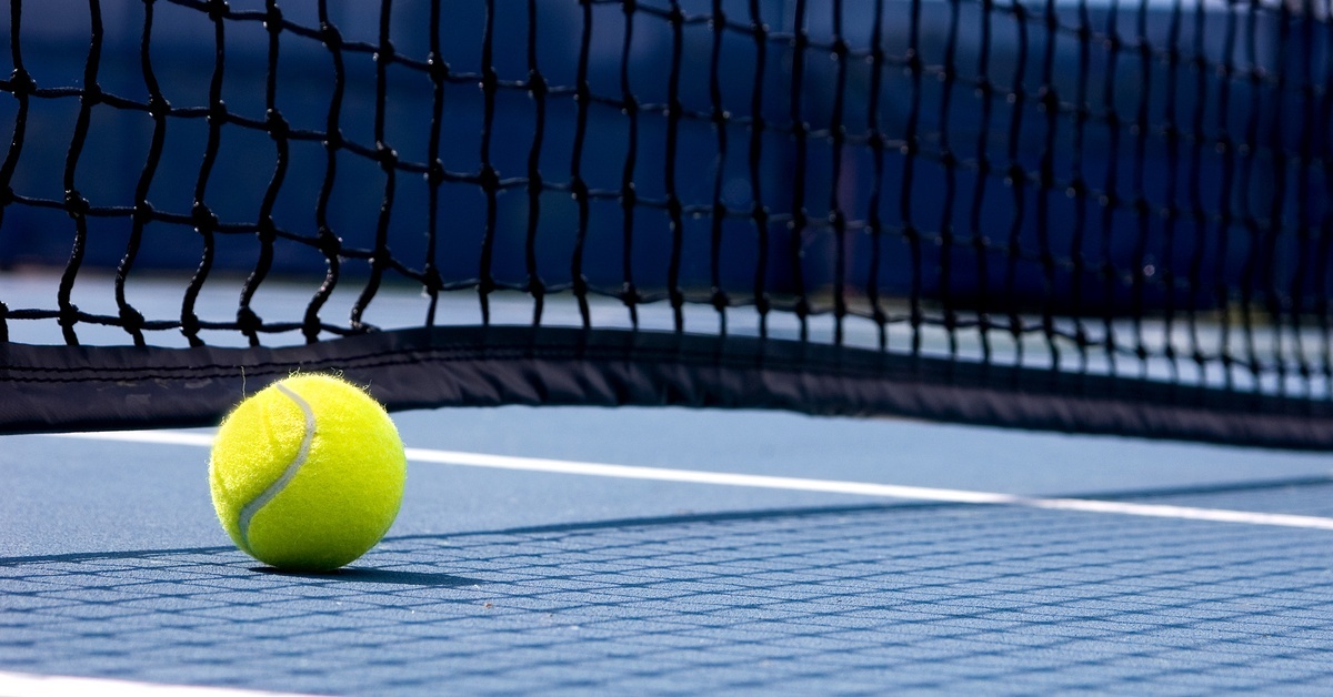 A traditional yellow tennis ball rests beneath a black tennis net, on a blue and green court during the day.
