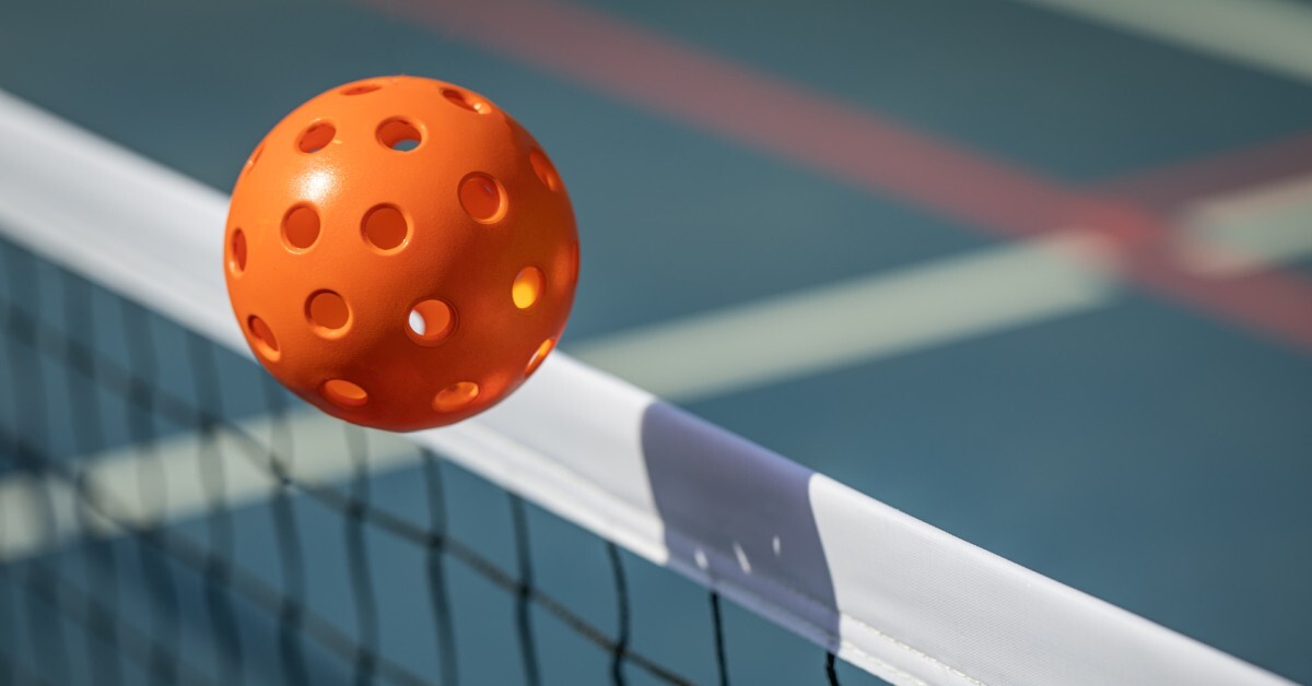 An orange pickleball hovers above a white net on a blue court, with blurred red and white lines in the background.