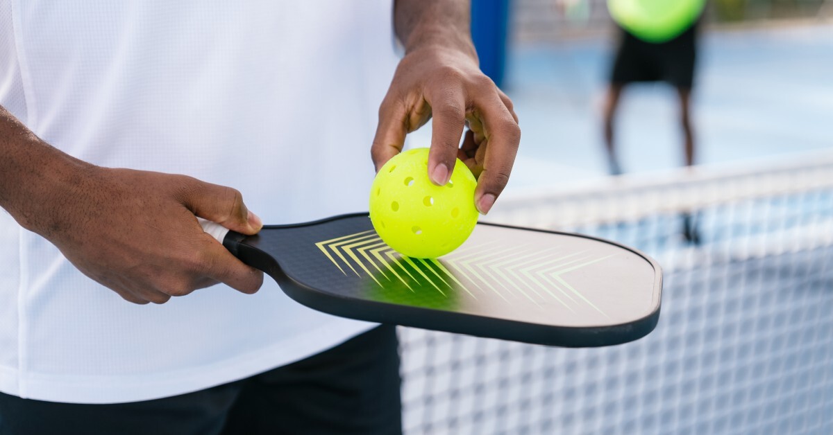  Hands hold a black pickleball paddle and a yellow ball on a court, with a blurred net and player in the background.