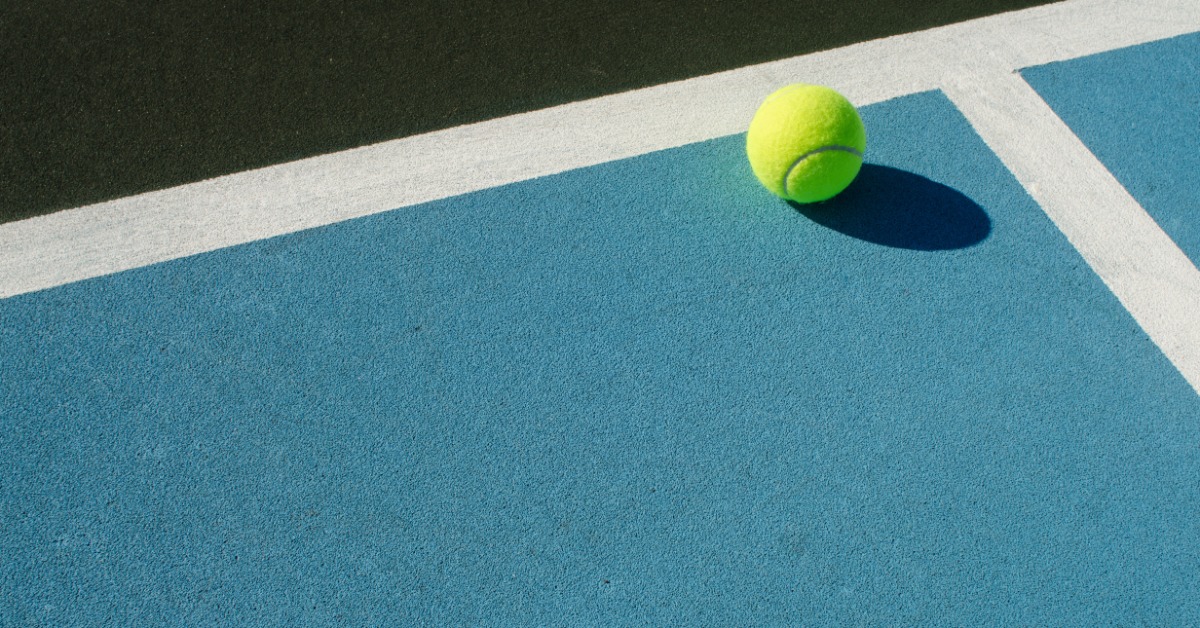 Tips for Organizing a Community Tennis Tournament Bright tennis ball resting near a white boundary line on a blue outdoor tennis court surface under direct sunlight.