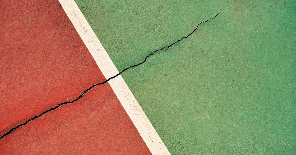 A crack running across a red and green tennis court surface near the boundary line, highlighting surface damage.