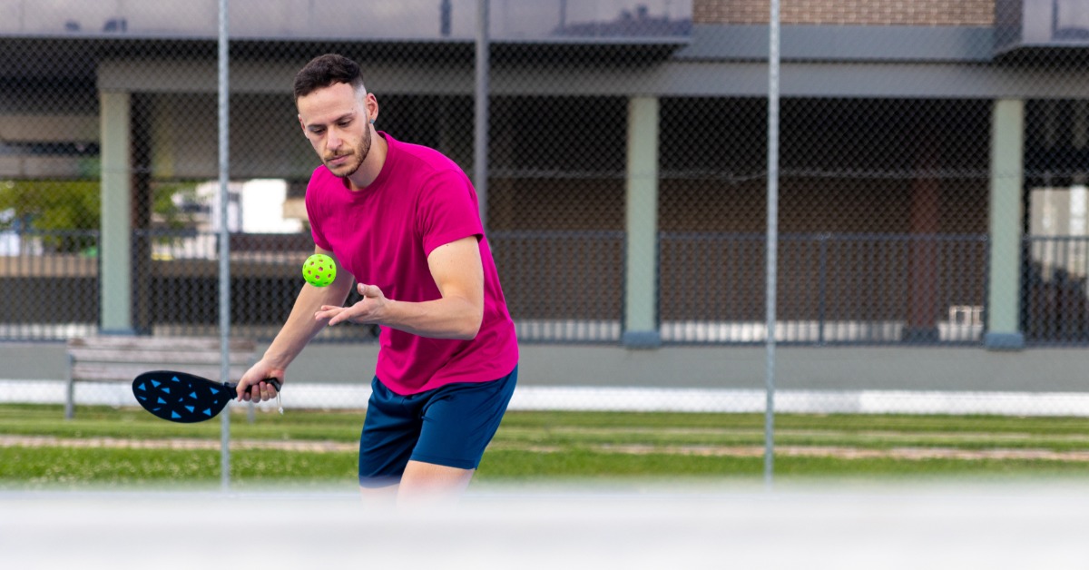 A man in a pink shirt prepares to serve a pickleball on an outdoor court with a net and fence in the background.