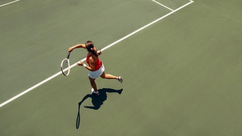 A woman holding a tennis racket on a green court, swinging near a yellow ball while viewed from directly overhead. A woman holding a tennis racket on a green court, swinging near a yellow ball while viewed from directly overhead.