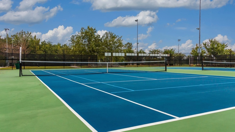An empty blue and green tennis court viewed from a corner, with white lines, a net, and trees beyond the fence. An empty blue and green tennis court viewed from a corner, with white lines, a net, and trees beyond the fence.