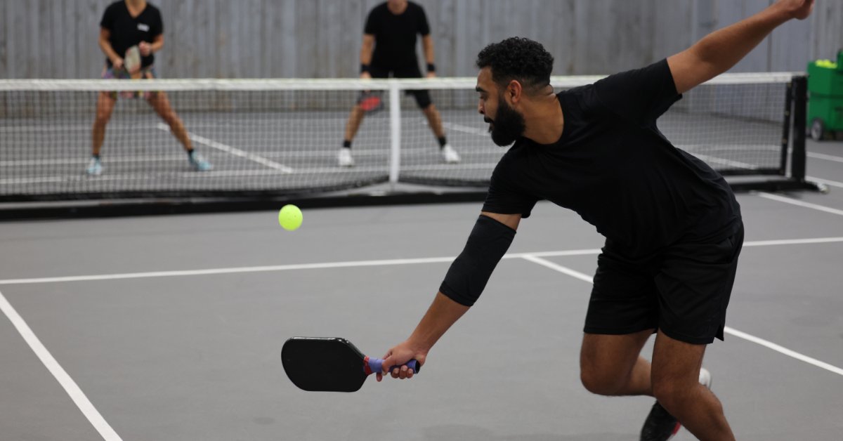 How To Organize a Pickleball Tournament in Your Community A player reaches down with a paddle toward a yellow ball on an indoor pickleball court while two players stand behind a net.
