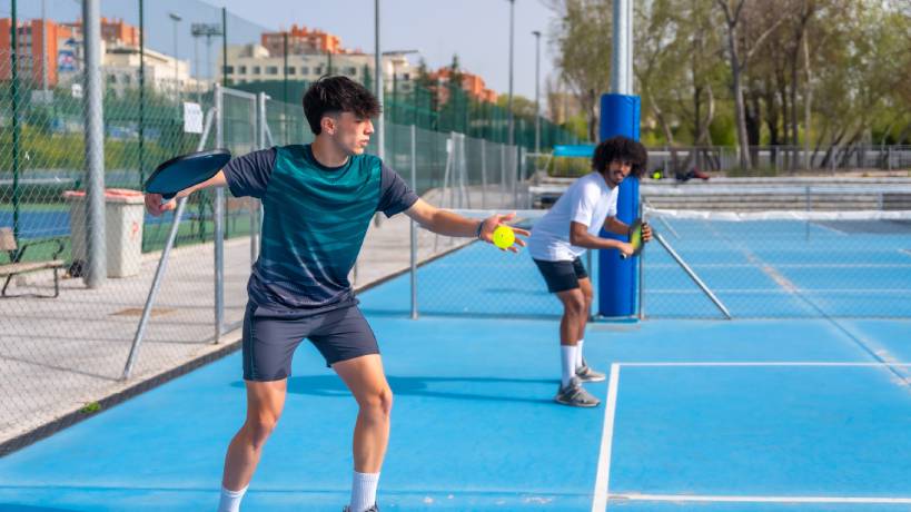 Two young men playing outdoor pickleball on the same team. The man on the right prepares to serve the ball.