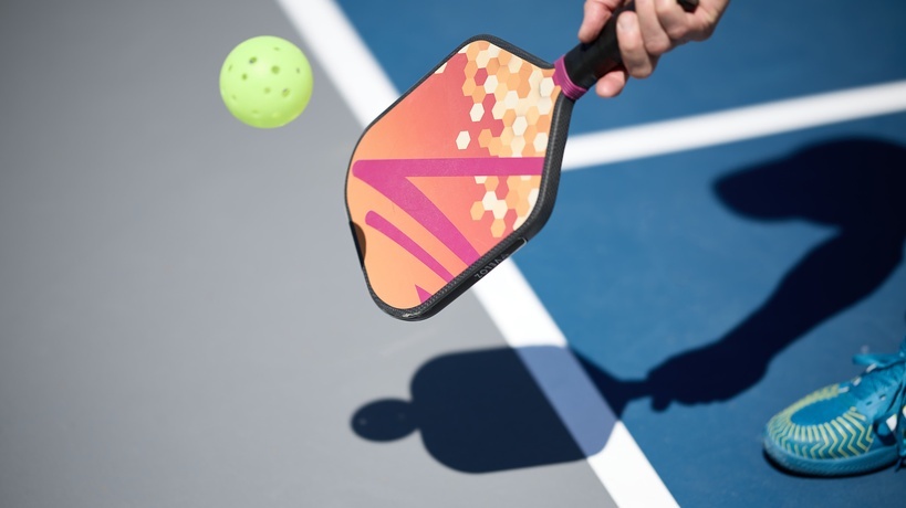 A hand holds a pickleball paddle with a unique graphic design. The paddle nears contact with a yellow pickleball. A hand holds a pickleball paddle with a unique graphic design. The paddle nears contact with a yellow pickleball.