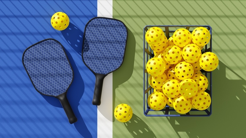 A basket contains a ton of yellow pickleballs. On the left, two yellow pickleballs surround two blue paddles.
