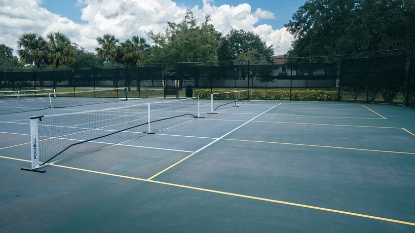 A pair of portable pickleball nets are on the blue outdoor tennis courts. Trees and blue, cloudy skies are in the background.