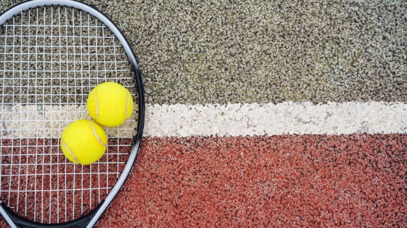 A racquet lays flat on a red and grey acrylic tennis court with white lines. Two yellow tennis balls are on top of the racquet.