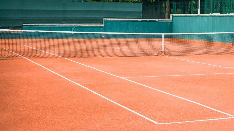An outdoor red clay tennis court has clean white lines, a tennis court net, and a green concrete backdrop.