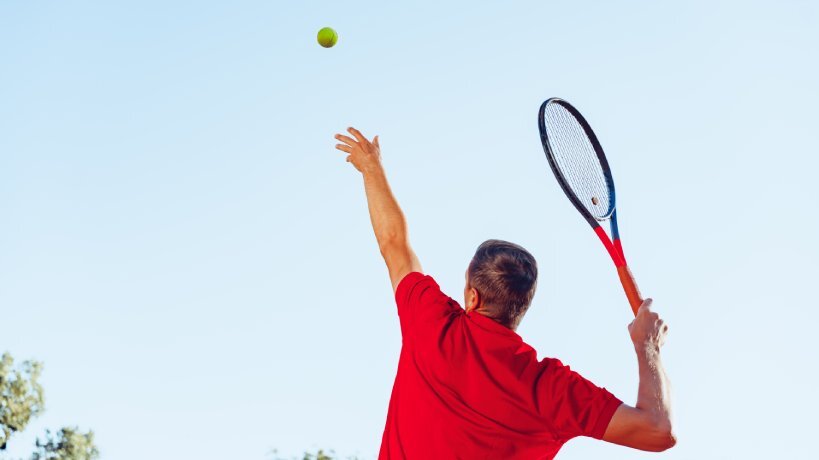 A man wearing a red shirt tosses a tennis ball with his left hand and prepares his racquet to serve with his right hand.