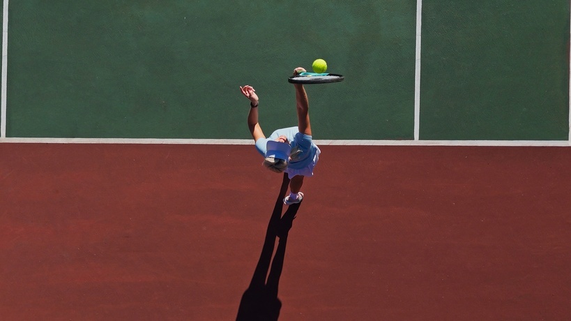A woman wearing a light blue tennis outfit hits a serve on a green hardcourt with a red backdrop.