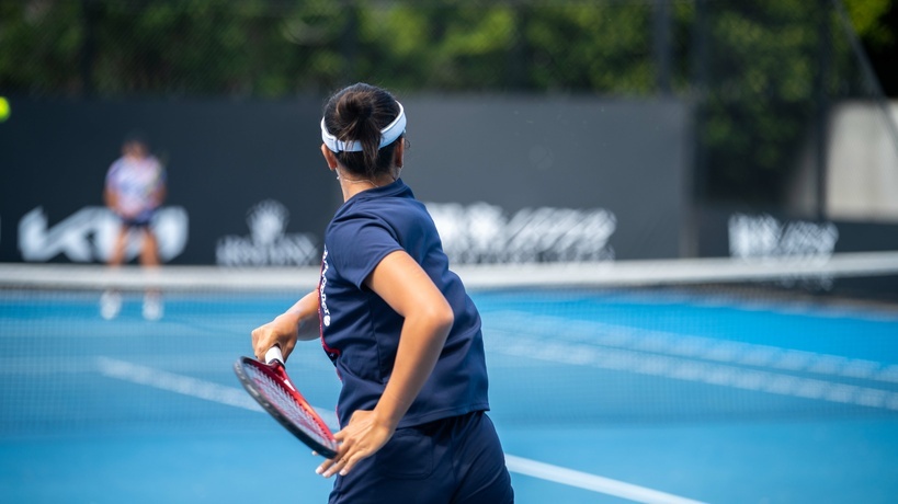 A young woman wearing a white visor, a navy shirt, and shorts hits an inside-out forehand on a hard court.