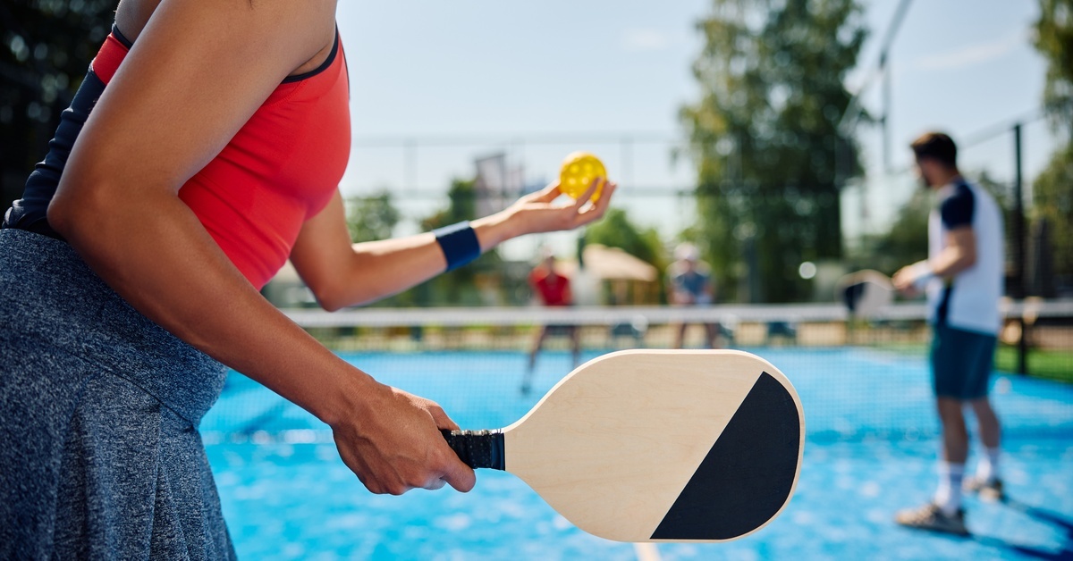 A woman wearing an orange top and grey skirt prepares to serve during a pickleball doubles match.