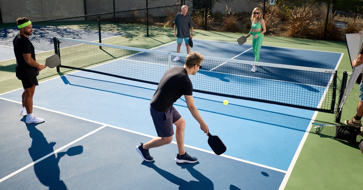 Three men and one woman play a pickleball doubles match on a blue court. One guy bends low and prepares to hit a dink shot.