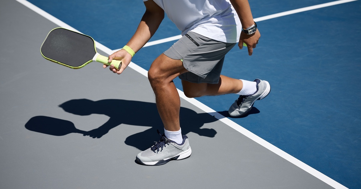 A pickleball athlete wearing a white shirt and grey athletic shorts holds a pickleball racquet and works on his footwork.