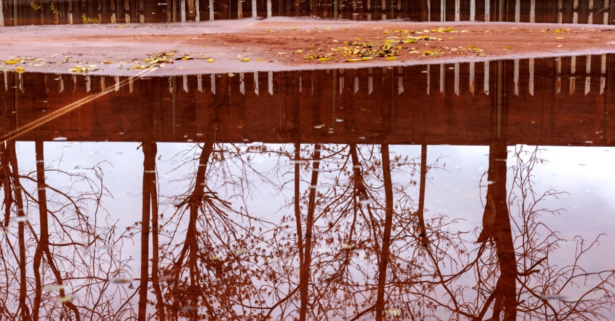 A wet red clay tennis court with puddles reflecting nearby trees after recent rainfall in an outdoor area.