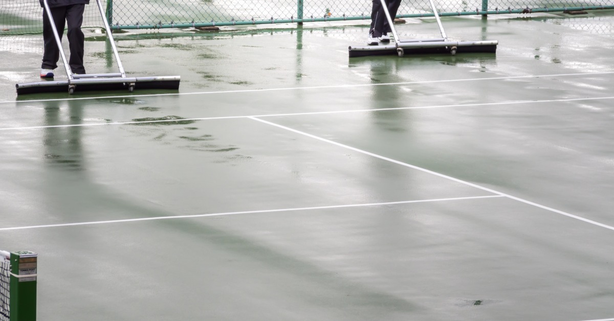 Workers using equipment to remove standing water from a tennis court after rainfall during the daytime.